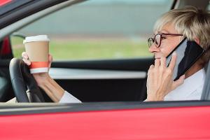 Reckless, smiling mature woman talking on the phone and holding a cup of coffee while driving a car