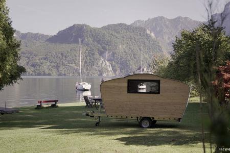 Der kleine Holz-Wohnwagen steht am Ufer eines Sees. Im Hintergrund segelt ein Boot vorbei und bewaldete Hügel säumen das Wasser.