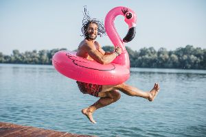 Mann mit pinkem Flamingo-Schwimmring springt vor Gewässer lachend in die Luft
