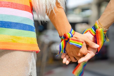 Zwei Menschen mit Regenbogenbändern am Armgelenkt fassen sich an den Händen