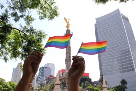 Wolkenkratzer im Hintergrund, davor ein Park mit einer Statue, vor der zwei Regenbogenfahnen gehalten werden.