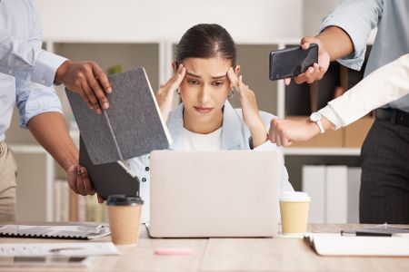 Frau sitzt gestresst am Tisch, weil ihr mehrere Menschen gleichzeitig Terminkalender, Telefone, Laptops, etc. reichen