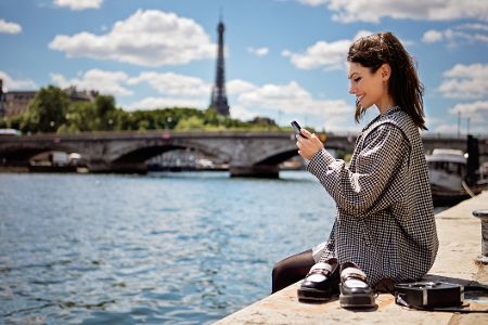 Frau sitzt an der Seine, im Hintergrund der Eiffelturm, hat ihre Schuhe ausgezogen und ihr Handy in der Hand