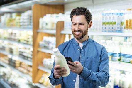 Lächelnder Mann im Lebensmittelgeschäft mit Smartphone zur Auswahl von Milchprodukten aus dem Kühlregal