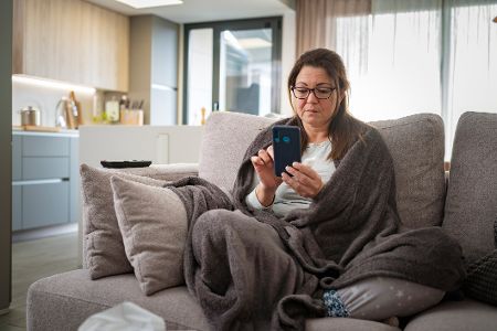Kranke Frau mit Wolldecke auf der Couch sitzt am Handy