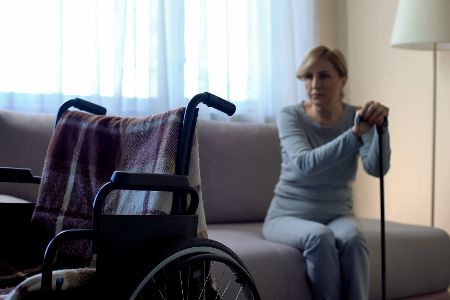 Frau sitzt auf einem Bett, in der Hand ein Gehstock, vor ihr ein Rollstuhl