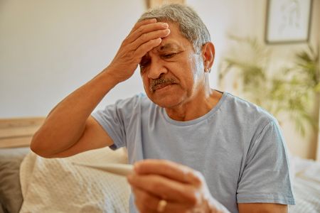 Mann hält sich die Stirn und hat Fieberthermometer in der Hand