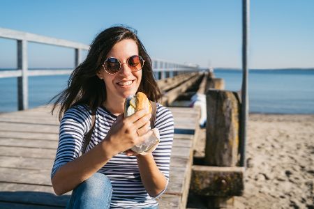 Frau sitzt am Strand auf einem Steg und isst ein Fischbrötchen