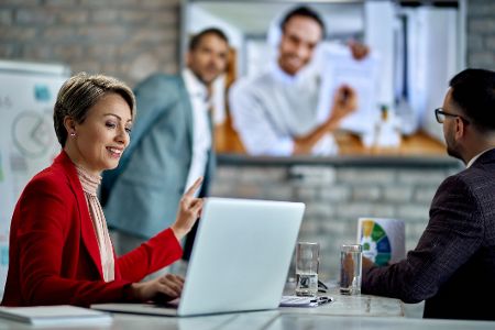 Frau am Laptop erklärt während eines Meetings mit mehreren Männern etwas