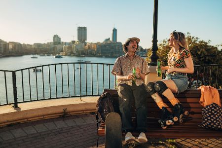 Zwei junge, hippe Leute sitzen mit Rollerblades am Ufer eines Flusses und trinken Bier