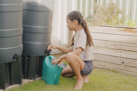 Kind holt mit Gießkanne Wasser aus mehreren Regentonnen