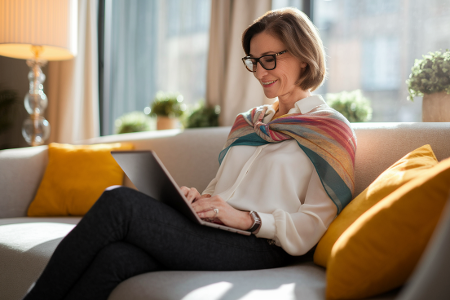 Eine Frau mittleren Alters sitzt entspannt auf einem Sofa und arbeitet lächelnd mit einem Laptop.