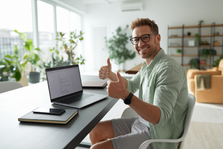 Mann sitzt in modernem Homeoffice am Laptop, zeigt Daumen hoch in die Kamera.