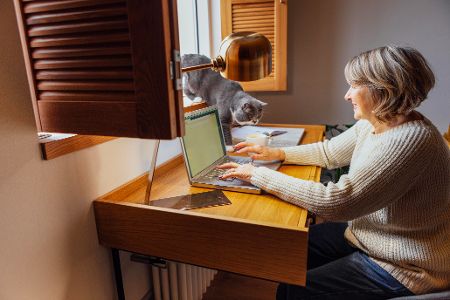 Ältere Frau sitzt am Computer, durch das offene Fenster klettert eine Katze zu ihr auf den Tisch