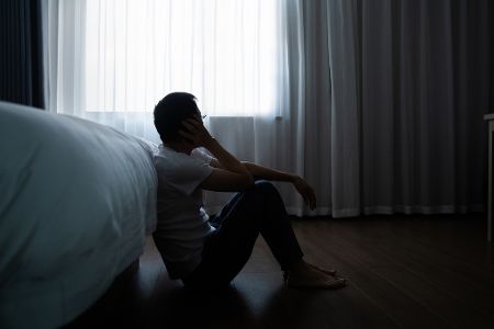 Mann sitzt in abgedunkeltem Raum an ein Bett gelehnt auf dem Fußboden