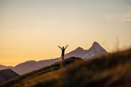 Mensch steht mit ausgebreiteten Armen auf einem Berggipfel im Sonnenuntergang
