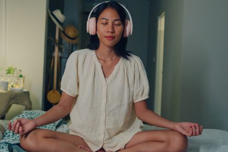 Junge Frau sitzt mit Kopfhörern auf einem Bett und meditiert