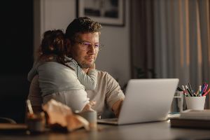 Mann sitzt im Homeoffice vor dem Laptop und hat ein kleines Baby auf dem Arm