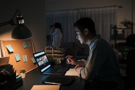 Mann arbeitet abends am Laptop, während die Frau im Hintergrund mit dem Kind spielt