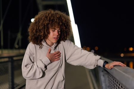 Junge Frau steht nachts an einer Brücke und fasst sich schmerzverzerrt an die Brust