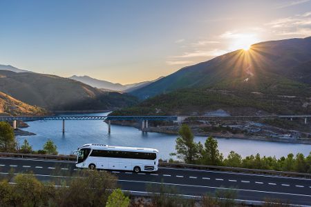 Bus fährt auf einer Straße vor einer Kulisse mit einem Fluss, einer Brücke und Bergen