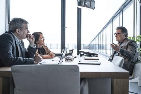 Drei Personen in einem modernen Büro bei einem Besprechungstermin an einem Konferenztisch. Zwei Personen sitzen auf der einen Seite, eine Person auf der anderen Seite und spricht gestikulierend. Laptops und Unterlagen liegen auf dem Tisch.