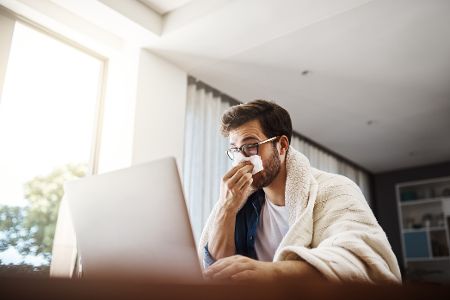 Mann sitzt mit Decke vor einem Laptop und putzt sich die Nase