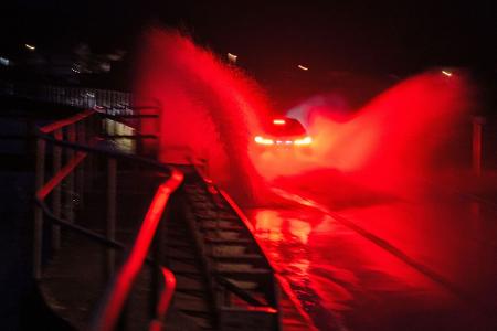 Wasser spritzt, während ein Auto über eine überschwemmte Straße zwischen Gleusdorf (Landkreis Haßberge, Unterfranken) und Busendorf (Landkreis Bamberg, Oberfranken) fährt.