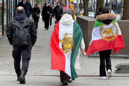 Menschen, die verschiedene ehemalige Flaggen des Iran umhaben, gehen durch die Münchner Innenstadt.