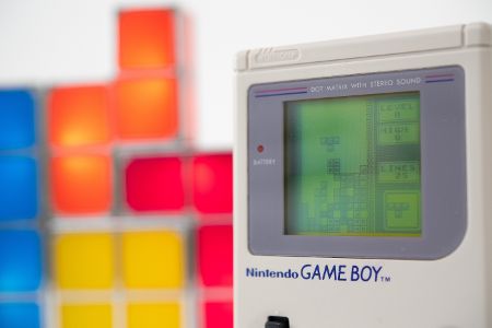 Gameboy mit einem laufenden Tetrisspiel steht vor einer Terisfigur