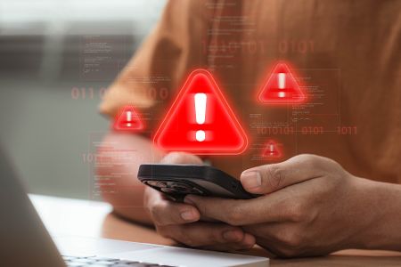 Mann vor einem Laptop sitzend hat ein Handy in der Hand, aus dem virtuelle Ausrufezeichen als Warnung aufsteigen