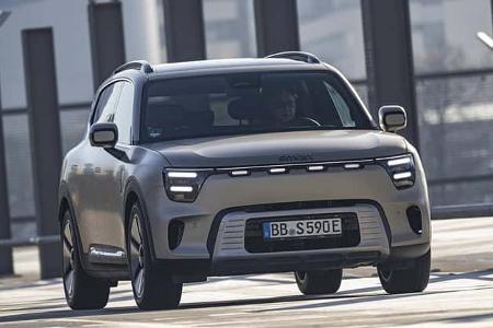 Das Bild zeigt einen Smart #5, ein modernes Elektro-SUV, frontal fahrend in urbaner Umgebung. Das Fahrzeug ist in silberner Lackierung gehalten und trägt ein deutsches Kennzeichen. Die Aufnahme entstand bei Tageslicht auf einer städtischen Straße mit architektonischem Hintergrund.