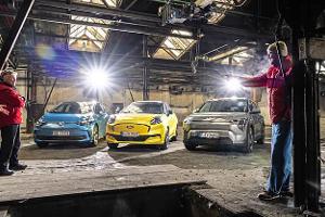 Drei Elektroautos – Ford Puma Gen-E, Kia EV3 und VW ID.3 – stehen nebeneinander in einer alten Industriehalle. Mehrere Personen in roten Jacken stehen um die Fahrzeuge, die Szene ist beleuchtet und vermittelt eine Test- oder Vergleichssituation.