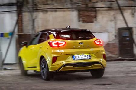 Ford Puma Gen-E Premium in gelber Lackierung, fotografiert von hinten in einer Industriehalle. Das Fahrzeug ist in Bewegung, die Rücklichter sind eingeschaltet. Die Aufnahme zeigt das Elektro-SUV in einer Indoor-Umgebung mit sichtbaren Hallenstrukturen.