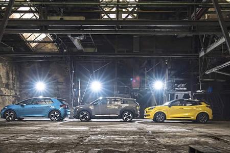 Drei Elektroautos – Ford Puma Gen-E, Kia EV3 und VW ID.3 – nebeneinander in einer Industriehalle, seitlich beleuchtet.