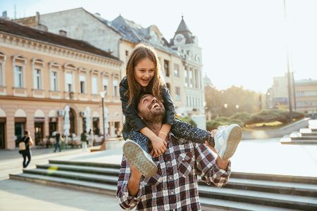 Vater steht auf einem Marktplatz in einer STadt und hat seine Tochter auf den Schultern