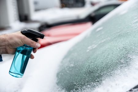 Hand mit Sprühflasche besprüht die vereiste Frontscheibe eines Autos