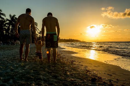 Zwei Männer mit einem kleinen Baby an der Hand laufen einen Strand entlang