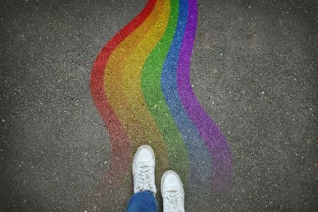 Zwei Beine mit weißen Turnschuhen stehen vor gewunden gemalten Pride-Farben auf Asphalt