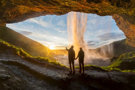 Zwei Personen stehen in einer Höhle hinter einem Wasserfall und schauen nach draußen