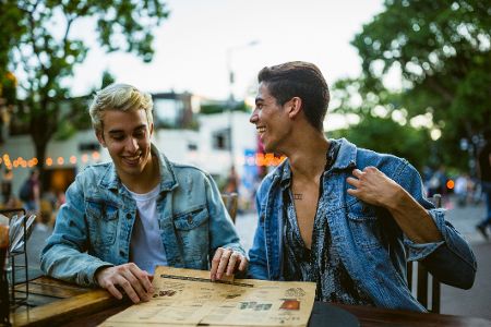 Zwei queere Männer lachen gemeinsam über einer Menükarte im Außenbereich eines Restaurants