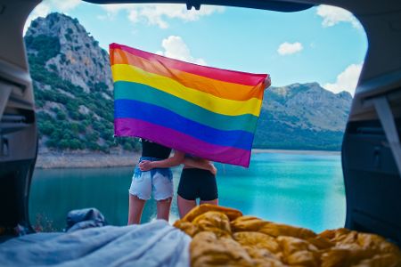 Zwei Frauen vor ihrem Zelt stehen vor einem See mit Gebirge im Hintergrund und halten eine Pride-Flagge
