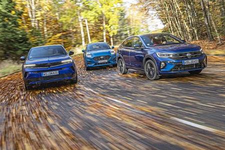 Drei kompakte SUV-Modelle – VW Taigo 1.5 TSI, Opel Mokka Hybrid und Ford Puma 1.0 EcoBoost – fahren nebeneinander auf einer herbstlichen Landstraße.