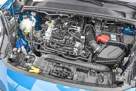 Detailaufnahme des Motorraums eines Ford Puma 1,0 EcoBoost. Der Dreizylinder-Turbobenziner ist mit allen Anbauteilen und Leitungen sichtbar. Die Aufnahme zeigt den Motorblock, Luftfilterkasten, Batterie und verschiedene technische Komponenten im geöffneten Motorraum eines blauen Fahrzeugs.