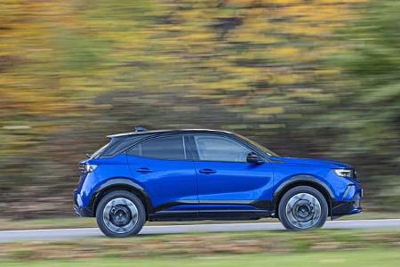 Opel Mokka Hybrid in blauer Lackierung fährt auf einer Straße vor herbstlicher Kulisse.