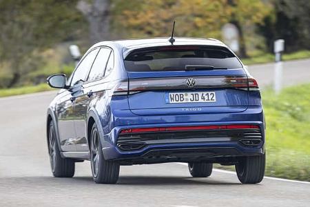 Das Bild zeigt einen VW Taigo 1,5 TSI auf einer Landstraße aus der Heckperspektive. Das Fahrzeug ist in blauer Lackierung zu sehen und fährt auf einer kurvigen Straße durch eine grüne, leicht bewaldete Landschaft.