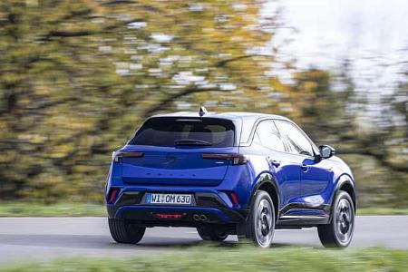 Opel Mokka Hybrid auf einer Landstraße, fotografiert von hinten in Fahrt, herbstliche Bäume im Hintergrund.