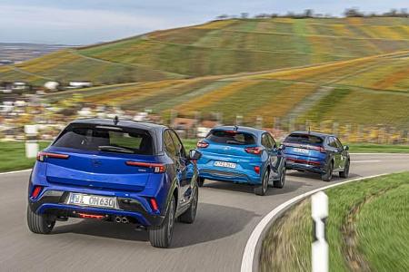 Drei kompakte SUV-Modelle – Opel Mokka, Ford Puma und VW Taigo – fahren in einer Gruppe auf einer kurvigen Landstraße durch eine hügelige Landschaft mit Weinbergen.