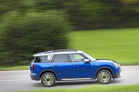 Seitenansicht eines blauen Mini Countryman C 2,0 auf einer Landstraße, fahrend vor grünem Hintergrund.