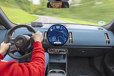 Innenraum des Mini Countryman C 2,0 während einer Fahrszene, Blick über die Schulter des Fahrers auf das Cockpit mit großem, rundem Zentraldisplay und moderner Instrumentierung. Der Fahrer trägt eine rote Jacke, die Straße ist durch die Windschutzscheibe sichtbar.